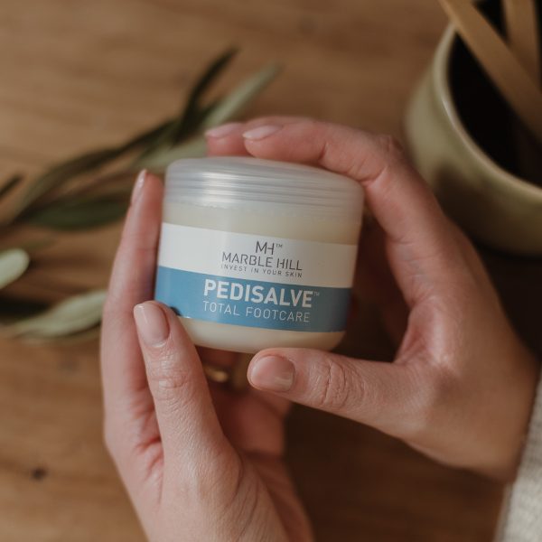 Hands holding Marble Hill Pedisalve Total Footcare product with wooden background